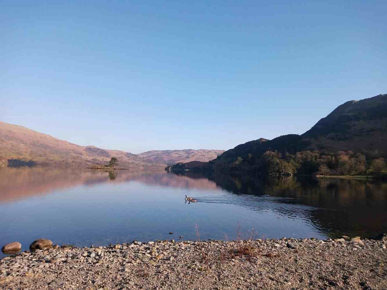 A stunning panoramic view of Ullswater in the Lake District, with calm waters reflecting the rolling hills and vibrant Spring landscape.